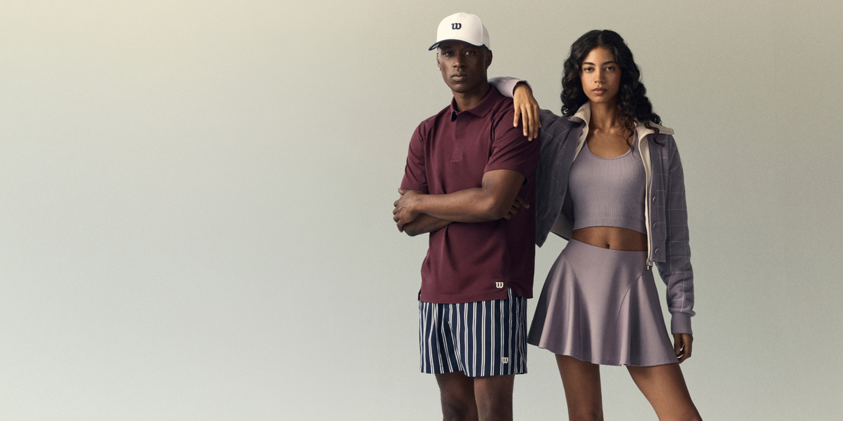Man in a maroon polo shirt and striped shorts with his arm around a woman in a lavender ribbed tank, skirt, and gray plaid jacket, standing against a sage green background