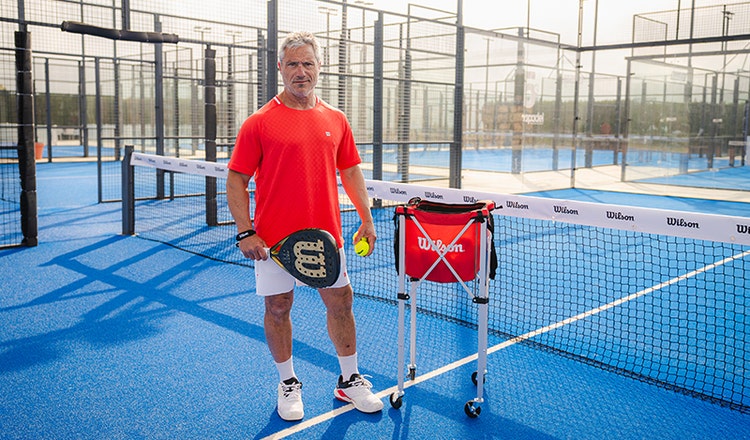 Padel coach Miguel Sciorilli on the court with a Wilson padel racket next to the net and a ball trolley.
