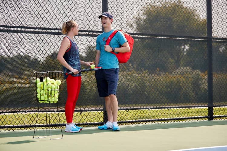 Two young tennis players on court with a bucket of tennis balls.
