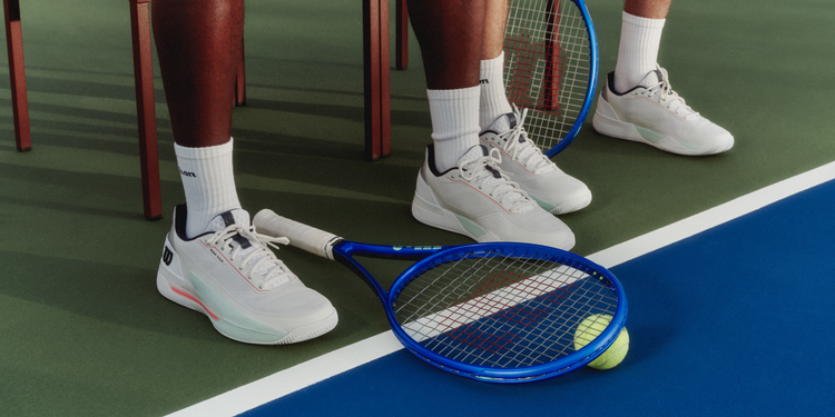Feet wearing the Wilson Rush 5 shoes on court and the Ultra tennis racket on the floor